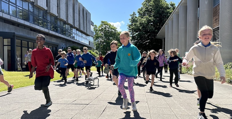 Grwp o blant yn ymweld a Campws Penglais Campus yn rhedeg gyda Smot, ein ci roboteg.