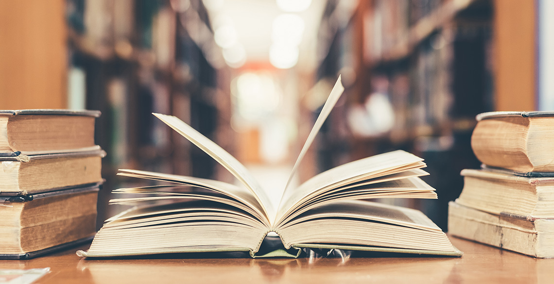 Open book on desk surrounded by other books, and library shelves in background